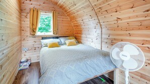 A cozy wooden room with a bed, a window with curtains, and a standing fan.