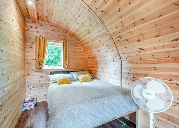 A cozy wooden room with a bed, a window with curtains, and a standing fan.