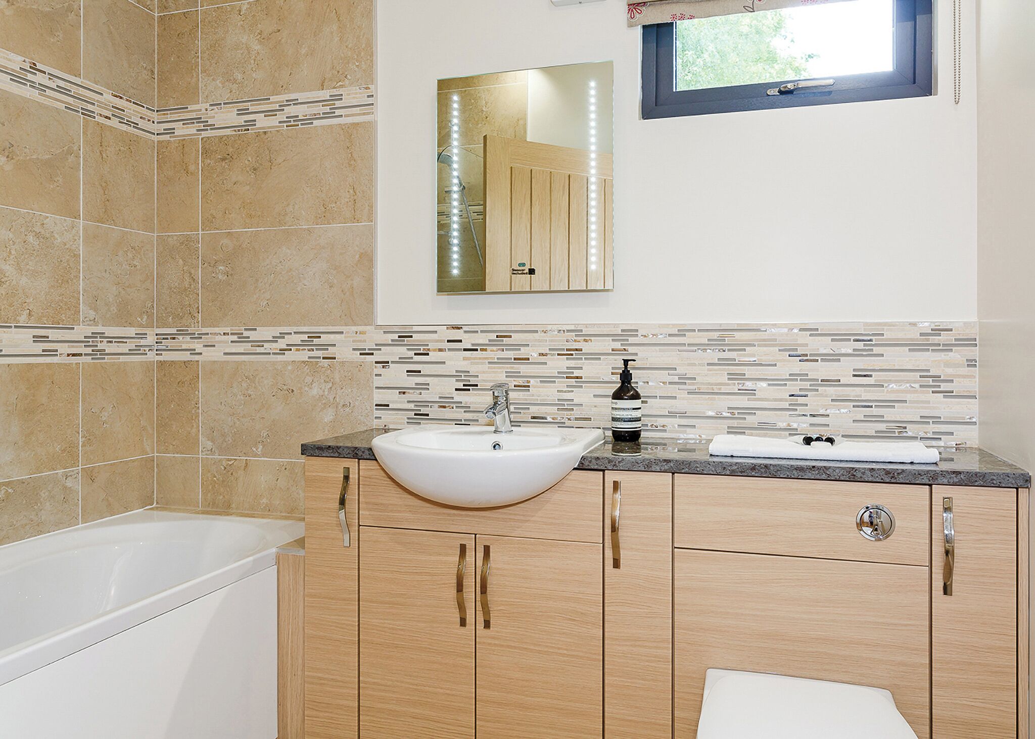 Un baño con bañera, lavamanos con dispensador de jabón, armarios de madera y salpicadero de azulejos.
