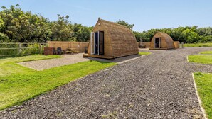 Two wooden cabins with open doors, a gravel path, and a grassy area with a wooden fence and trees in the background.