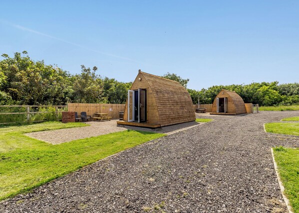 Two wooden cabins with open doors, a gravel path, and a grassy area with a wooden fence and trees in the background.