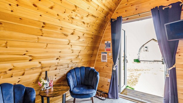 A wooden interior with a round table, two chairs, a clock, and a door leading outside.