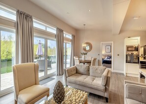 A spacious living room with a sofa, armchair, and dining table. There is a balcony with a view of greenery.