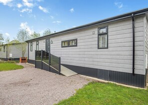 Un bâtiment d’un seul étage, avec une allée en gravier, une terrasse en bois et une petite cheminée en briques.