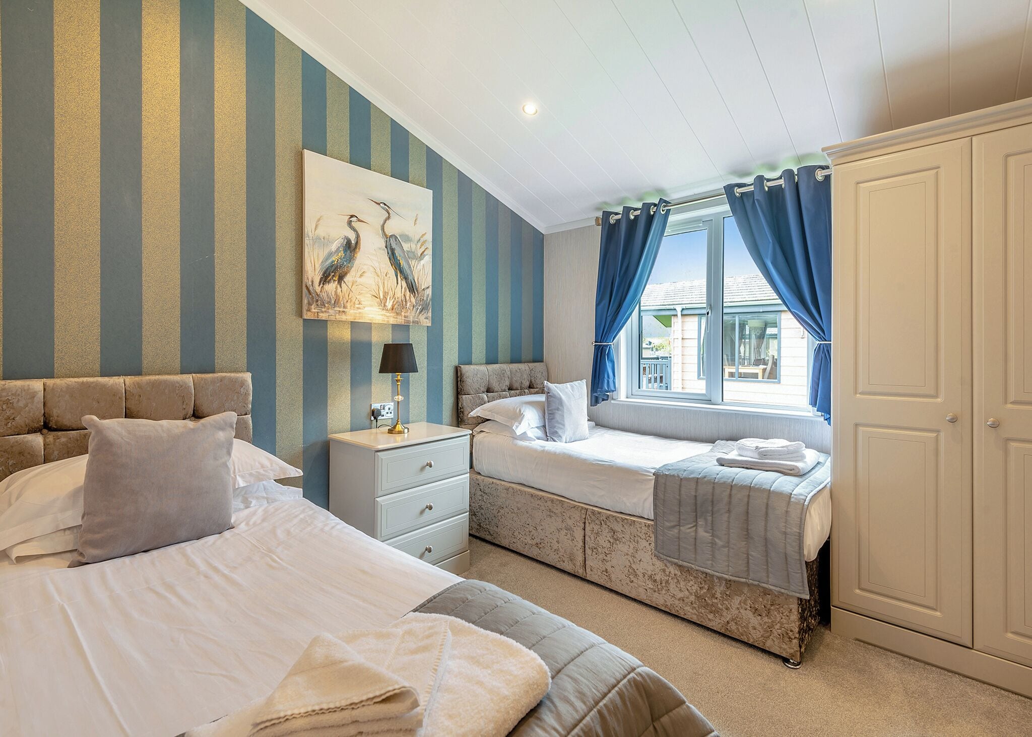 Une chambre d’hôtel avec deux lits, une table de chevet, une lampe, un tableau et une fenêtre avec des rideaux.