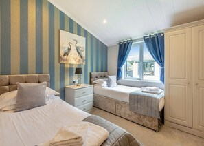 A hotel room with two beds, a nightstand, a lamp, a painting, and a window with curtains. - New in 2018. Modern and luxurious lodge with lakeside view. (Woodhall Spa)