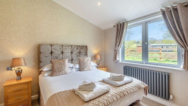 A bedroom with a large bed, two bedside tables with lamps, a window with curtains, and a radiator. - New in 2018. Modern and luxurious lodge with lakeside view. (Woodhall Spa)