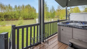 A covered outdoor deck with a hot tub, wooden flooring, and a view of a lake and trees. - New in 2018. Modern and luxurious lodge with lakeside view. (Woodhall Spa)