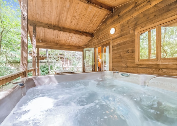 Una cabaña de madera con jacuzzi, ventanas amplias y vistas al bosque circundante.