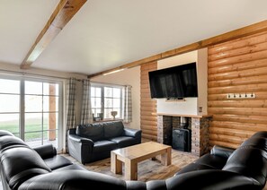Interior de una cabaña de madera con chimenea, sofá de cuero, mesa de centro de madera y televisor montado.