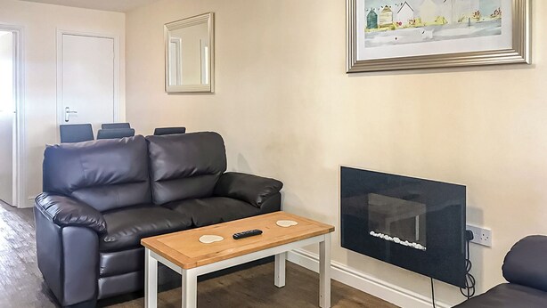 A living room with a black leather sofa, a wooden coffee table, and a flat-screen TV mounted on the wall. - Merryfield & Sandfields Apartment (Chapel St Leonards)
