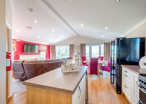 A modern kitchen with a central island, a dining area with red chairs, and a living area with a red wall and a flat-screen TV.
