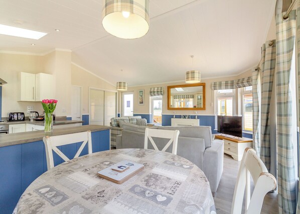 A modern kitchen with white cabinets, a dining table with a newspaper-patterned tablecloth, and a living area with a sofa and TV.