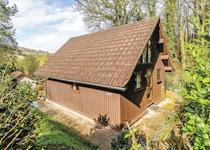 A wooden cabin with a sloped roof, surrounded by greenery and a clear sky.
