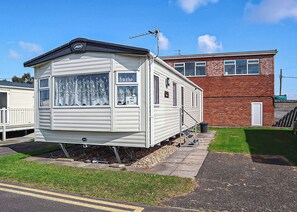 Une maison mobile équipée d’une antenne parabolique, d’un petit porche et, au fond, d’un bâtiment en briques.