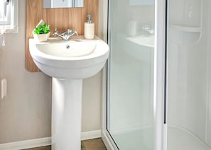 Ein Badezimmer mit einem weißen Standwaschbecken, einer Wandverkleidung aus Holz, einer kleinen Topfpflanze, einem Seifenspender und einer Duschkabine aus Glas.