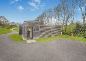 A small, modern building with a glass door, situated on a paved path with a grassy area and trees in the background.