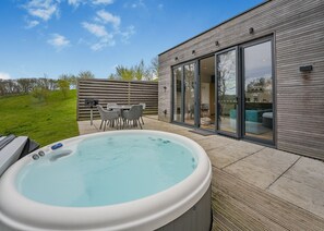 A hot tub with a patio, a dining area with chairs, and a modern building with large glass doors. - Woodland View Retreat (Pet) (Lewes)