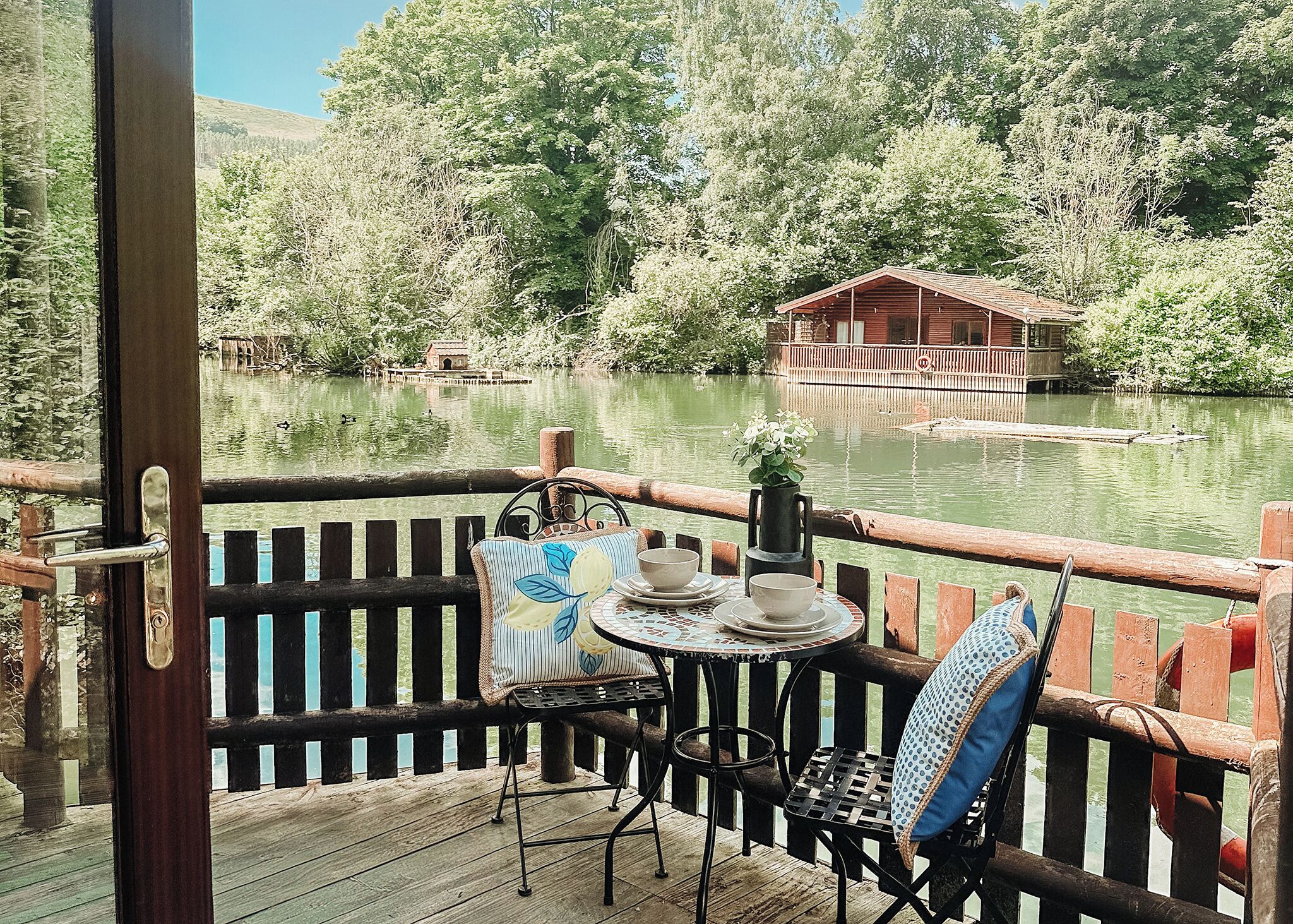 Une terrasse en bois avec une table et des chaises, donnant sur un lac paisible, avec un chalet en bois en arrière-plan.