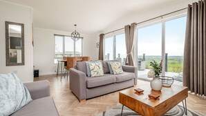 A spacious living room with a grey sofa, a wooden coffee table, and a dining area with a table and chairs. Large windows offer a view of the countryside.