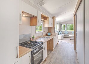 Una cocina moderna para vehículos recreativos con estufa, pileta y horno, integrada a un espacio de estar con sofá y ventanas amplias.