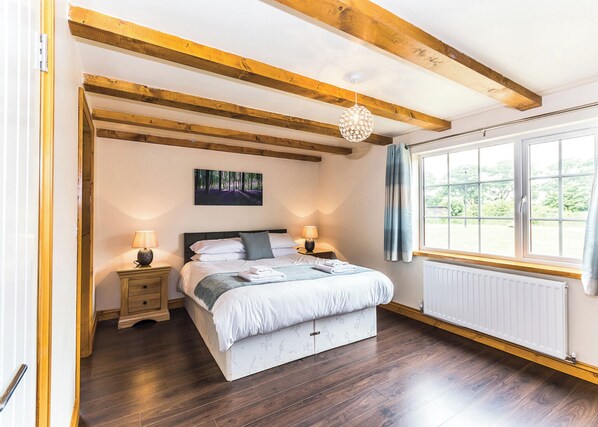 Une chambre avec un plafond en bois, un lit avec une literie blanche, deux tables de chevet avec des lampes, une fenêtre avec des rideaux et un radiateur.