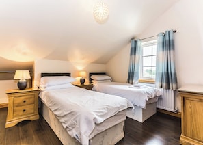 Ein Schlafzimmer mit zwei Betten, einem Nachttisch mit Lampe, einem Fenster mit Vorhängen und einem Holzboden.