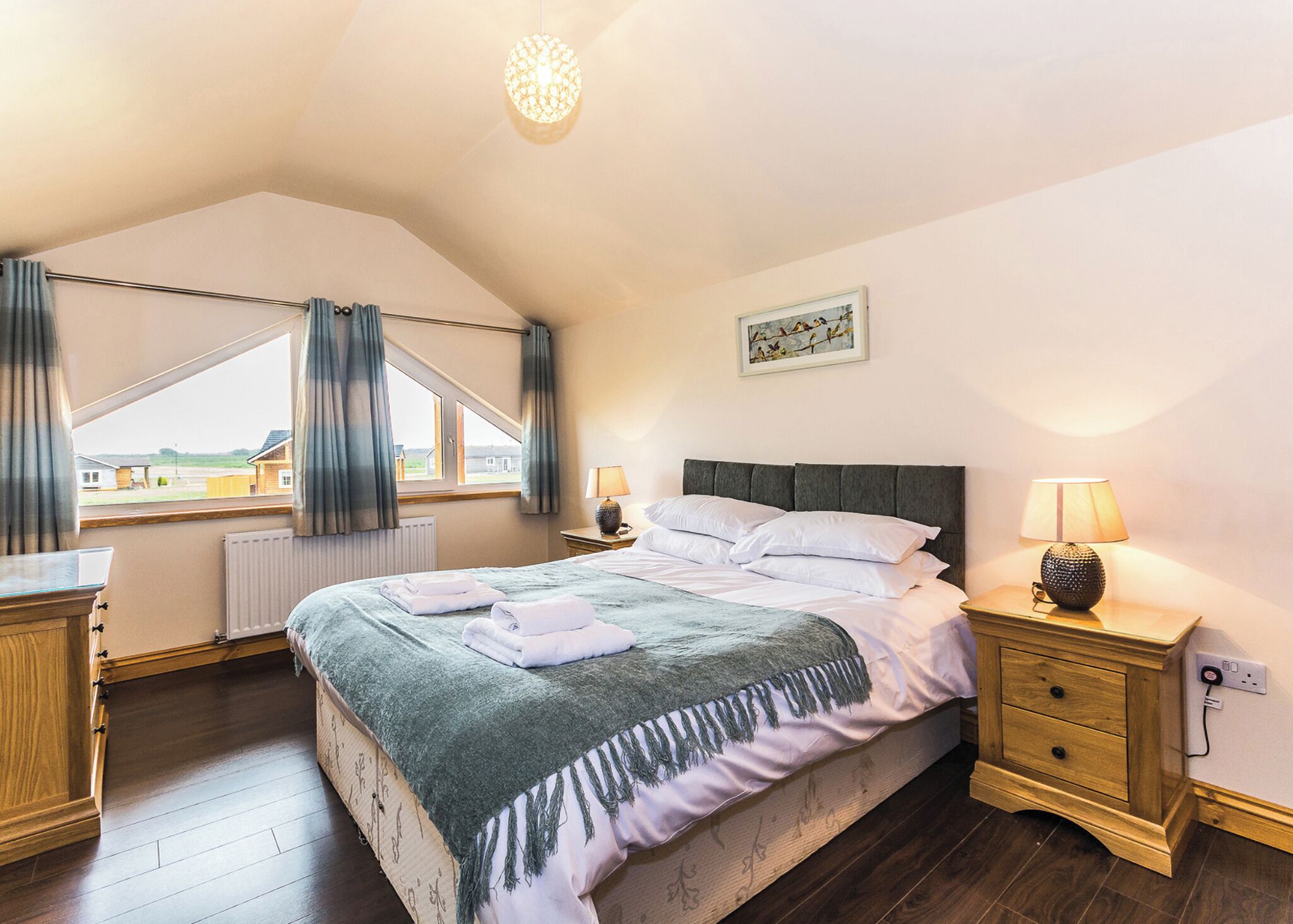 A bedroom with a bed, bedside tables, a lamp, a window with curtains, a radiator, and a painting on the wall.