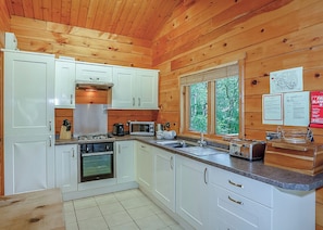 A wooden cabin kitchen with white cabinets, a stove, microwave, sink, and a window with a view of trees. - 2 bedroom accommodation in Freehay, Cheadle (Freehay, Cheadle)