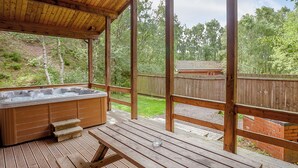 Une terrasse en bois avec un jacuzzi, une table de pique-nique et une vue sur une cour clôturée bordée d’arbres.
