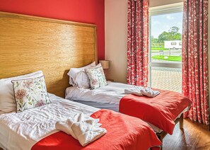 A hotel room with two beds, red and white bedding, wooden headboard, and a view of a green landscape through the window.