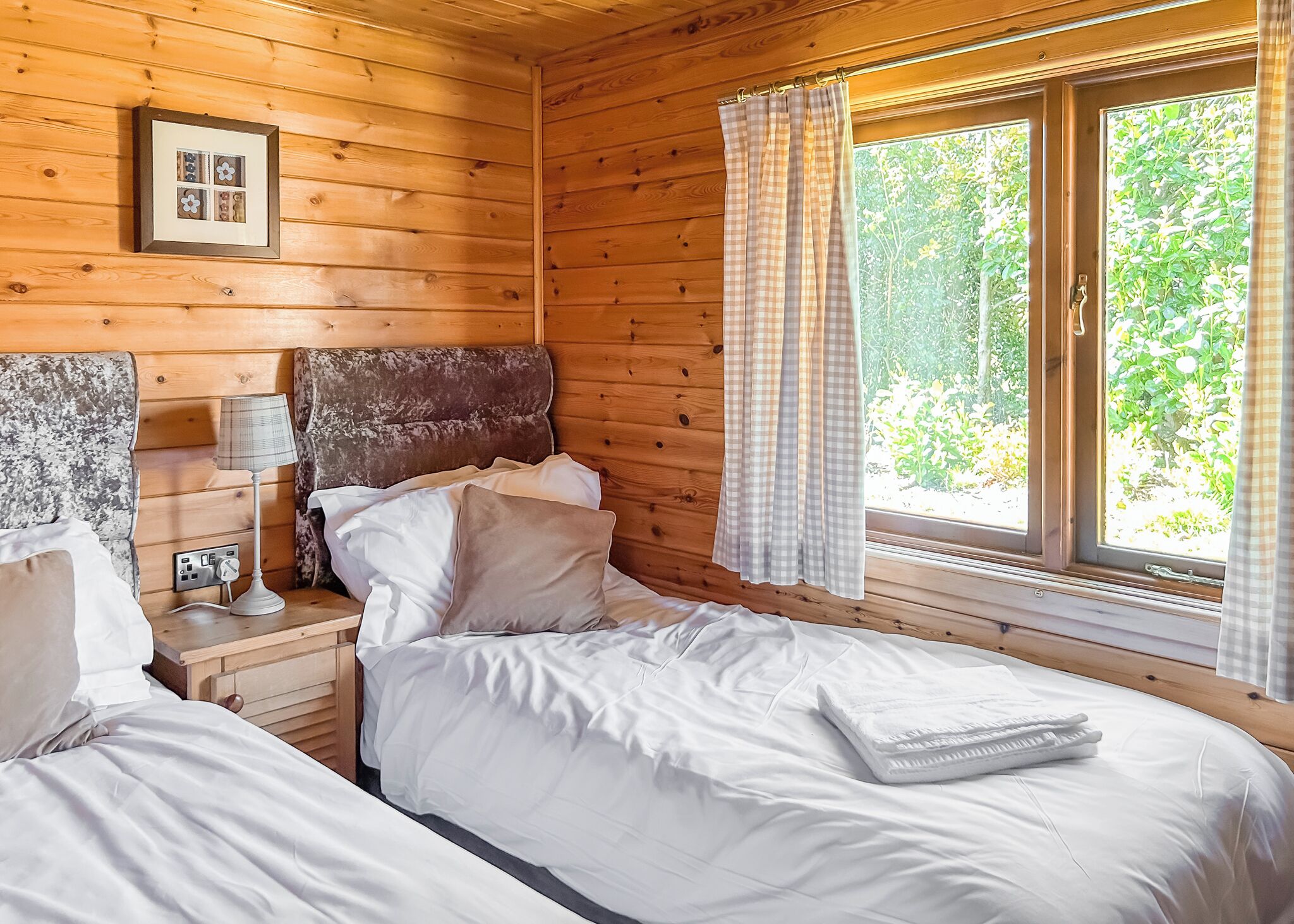 A wooden cabin bedroom with a bed, bedside table, lamp, and window with curtains.