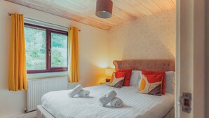 A neatly made bed with white linens and towels, a window with yellow curtains, and a wooden ceiling. - Woodpecker Lakeside Lodge (Caerwys, Flintshire)