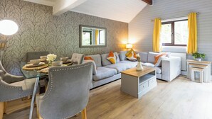 A living room with a sofa, chairs, a coffee table, and a dining table set with bowls and flowers. - Woodpecker Lakeside Lodge (Caerwys, Flintshire)