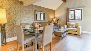 A modern living room with a dining area, a sofa, a coffee table, a lamp, and a window. - Woodpecker Lakeside Lodge (Caerwys, Flintshire)