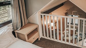 A bedroom with a bed, a wooden nightstand, a white crib, and a window with curtains. - Woodpecker Lakeside Lodge (Caerwys, Flintshire)