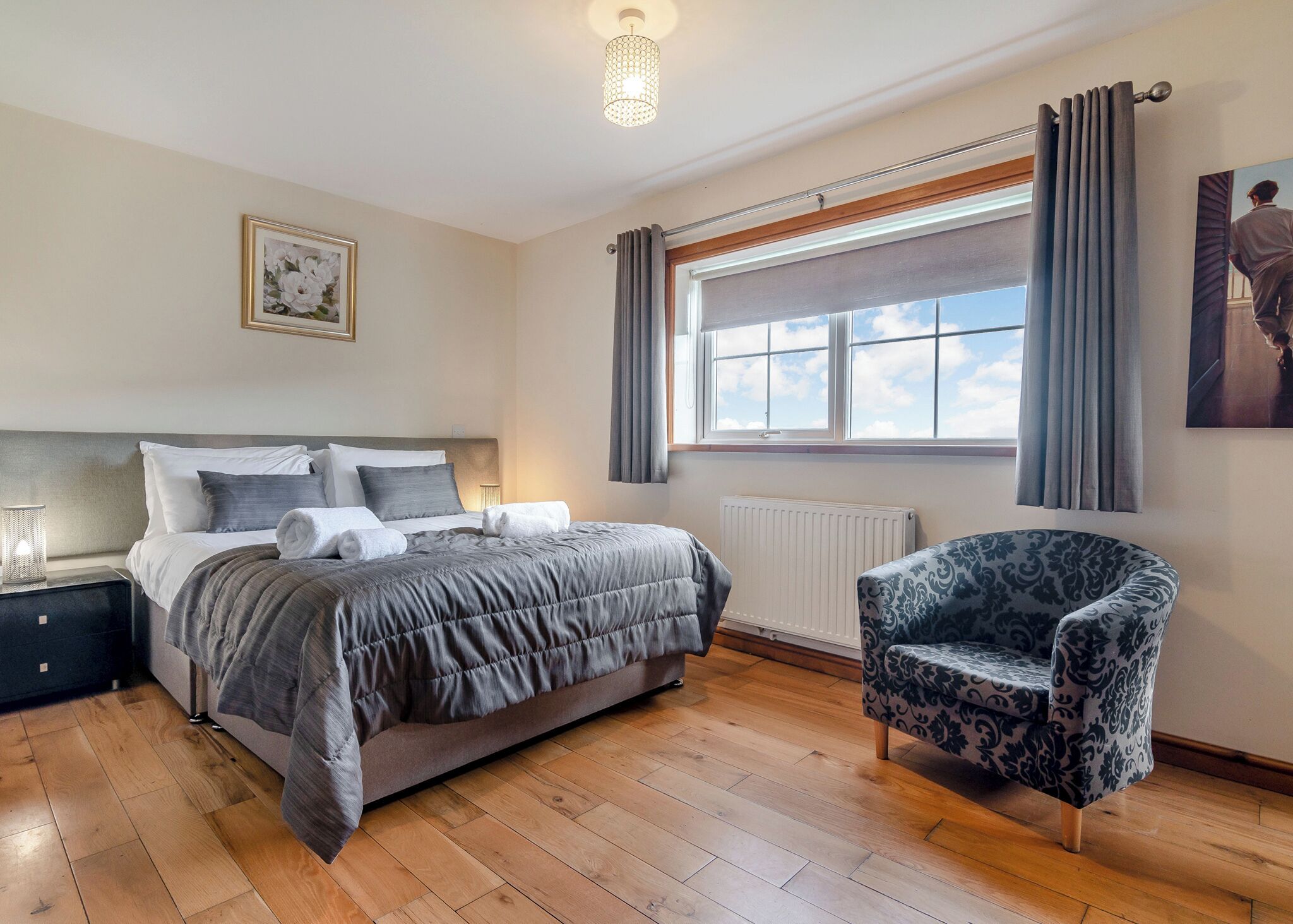 A bedroom with a bed, bedside table, lamp, chair, window, curtains, radiator, and a framed picture on the wall.