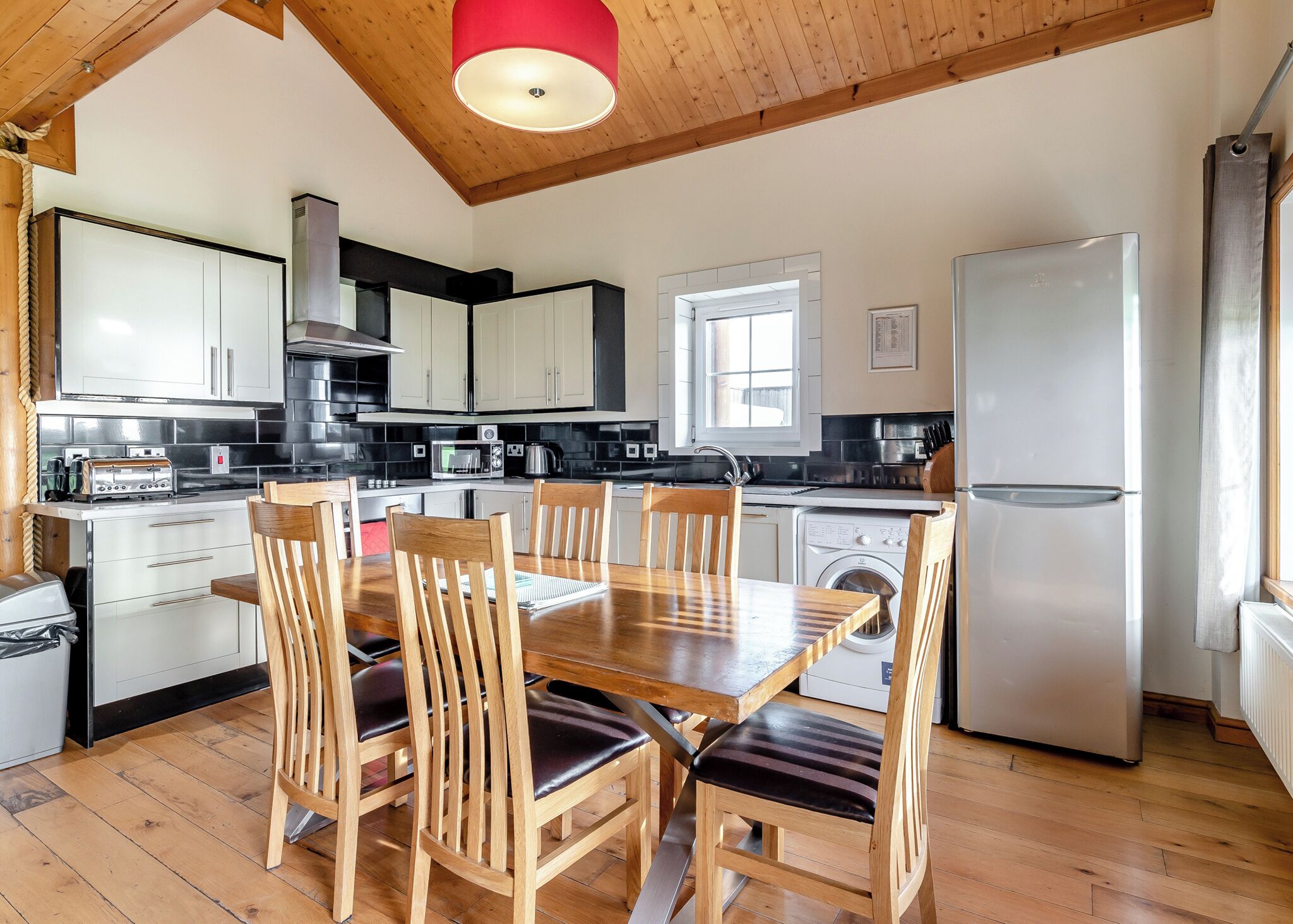A modern kitchen with wooden cabinets, a dining table with chairs, a washing machine, and a refrigerator.
