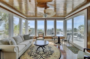 A spacious room with a large window, a ceiling fan, a sofa, a coffee table, and a dining area with chairs. - Oceanfront! Pool, Hot Tub, Walk to Town, Pets ok w/fee - 910 Ocean (Isle Of Palms)