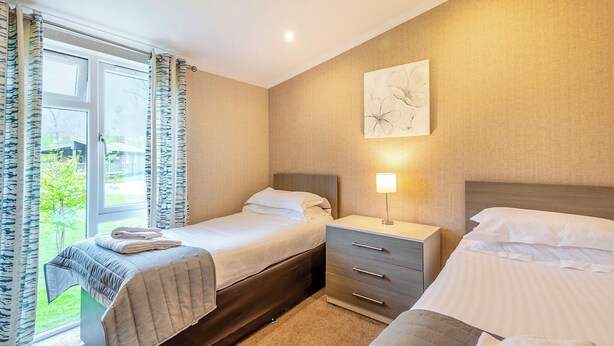 A hotel room with two beds, a bedside table with a lamp, a window with curtains, and a wall-mounted artwork. - Lakeside 2 Pet Friendly (Woodhall Spa)