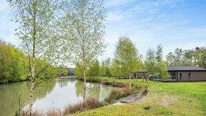 A serene lakeside setting with a small dock, grassy areas, and a building in the background. - Lakeside 2 Pet Friendly (Woodhall Spa)