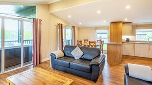 A spacious living area with a black leather sofa, wooden flooring, and a sliding glass door leading to a balcony. - Lakeside 2 Pet Friendly (Woodhall Spa)
