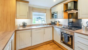 A modern kitchen with wooden cabinets, a stainless steel stove, a microwave, and a window with a view of a house. - Lakeside 2 Pet Friendly (Woodhall Spa)