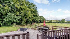 Une terrasse avec des sièges extérieurs, un parasol rouge et une vue sur un champ verdoyant, avec des arbres et une colline au loin.