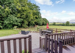 A deck with outdoor seating, a red umbrella, and a view of a grassy field with trees and a distant hill.