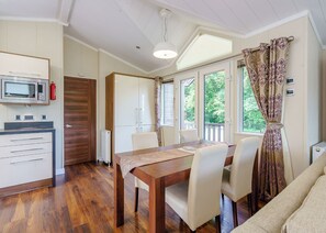 A dining area with a wooden table and chairs, a microwave, and a view of greenery through large windows.