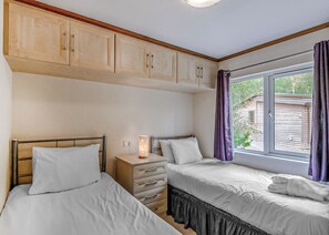 Un dormitorio con dos camas, armarios de madera arriba, una mesita de noche con lámpara y una ventana con cortinas.