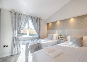 A modern bedroom with two beds, a large window with curtains, a stone-textured wall, and a bedside table with a vase.