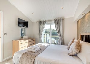 A bedroom with a bed, a TV mounted on the wall, a wooden dresser, and a window with curtains.
