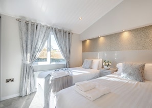 A modern bedroom with two beds, a large window with curtains, a stone-textured wall, and a bedside table with a vase.